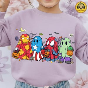 May include: A light purple crewneck sweatshirt featuring a graphic print of four characters dressed as ghosts for Halloween. The characters resemble Iron Man, Captain America, Spider-Man, and a green monster, each holding a small orange pumpkin bucket.