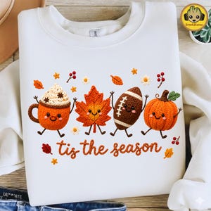 Pode incluir: Uma camisola branca de gola redonda com uma impressão gráfica de quatro personagens antropomórficas: uma caneca de chocolate quente, uma folha de ácer, uma bola de futebol americano e uma abóbora. O texto 'tis the season' está impresso a cor de laranja.