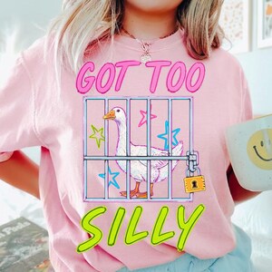K&ouml;nnte beinhalten: Ein hellrosa T-Shirt mit dem Text "GOT TOO SILLY" in leuchtendem Pink und Gr&uuml;n. Die Grafik zeigt eine wei&szlig;e Gans in einem K&auml;fig mit Sternen und einem Vorh&auml;ngeschloss. Die Person h&auml;lt eine Tasse mit einem Smiley.