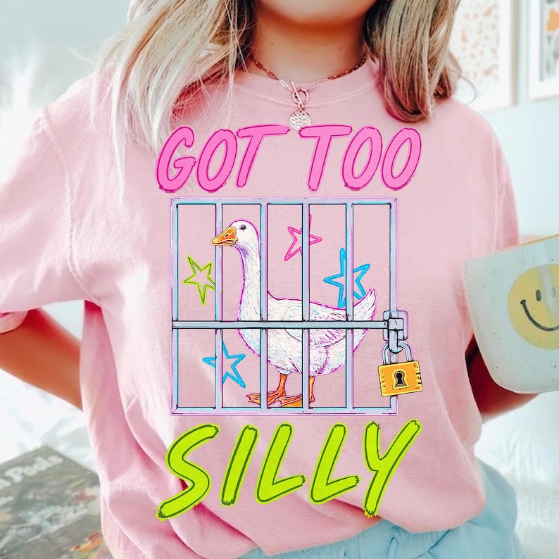 K&ouml;nnte beinhalten: Ein hellrosa T-Shirt mit dem Text "GOT TOO SILLY" in leuchtendem Pink und Gr&uuml;n. Die Grafik zeigt eine wei&szlig;e Gans in einem K&auml;fig mit Sternen und einem Vorh&auml;ngeschloss. Die Person h&auml;lt eine Tasse mit einem Smiley.