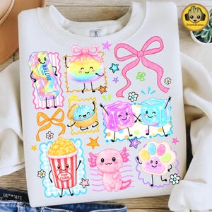 Könnte beinhalten: Ein weißes Sweatshirt mit einem bunten Grafikdruck verschiedener Zeichentrickfiguren, darunter ein Stück Butter, ein regenbogenfarbener Beutel, eine rosa Schleife, zwei Eiswürfel, ein Popcorn-Eimer, ein Axolotl und ein Pfotenabdruck.