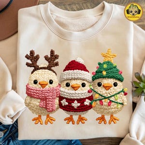May include: A cream-colored sweatshirt featuring three embroidered chicks dressed for the holidays. One chick wears reindeer antlers and a pink scarf, another a Santa hat and sweater, and the last a Christmas tree hat and lights.