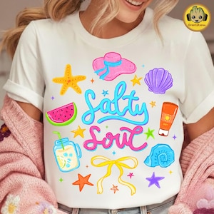 May include: White t-shirt with a colorful beach-themed graphic. The design includes a pink hat, watermelon slice, starfish, seashell, and the words "Salty Soul" in a playful font. A fun summer tee.