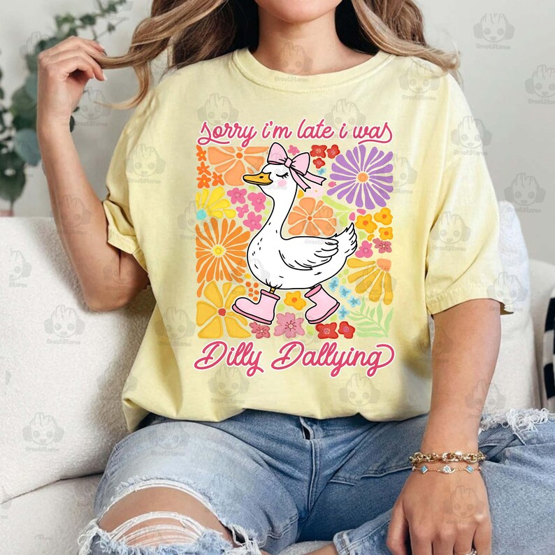 Op de afbeelding: Een geel T-shirt met een witte eend die roze laarzen en een roze strik draagt. De eend is omringd door kleurrijke bloemen. De tekst "Sorry, ik ben te laat, ik was aan het dralen" staat op het shirt gedrukt.