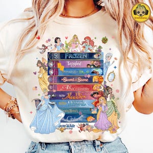 Puede incluir: Camiseta color crema con una pila de cintas VHS de Disney con títulos como "Frozen", "Tangled" y "Aladdin". Las princesas de Disney están representadas encima y alrededor de las cintas. La camiseta tiene cuello redondo y mangas cortas.
