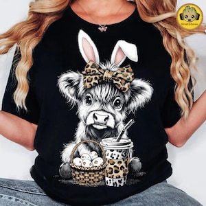 May include: Black t-shirt featuring a whimsical illustration of a Highland cow with bunny ears and a leopard print bow. The cow is holding a coffee cup and a basket of eggs. A fun Easter-themed design.
