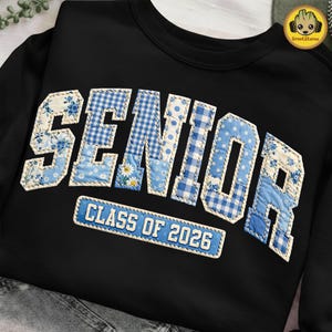 以下が含まれることがあります： 黒色のスウェットシャツ。パッチワーク文字で「SENIOR」と書かれており、青と白の花柄、水玉模様、ギンガムチェック柄が特徴です。「CLASS OF 2026」と書かれた長方形のパッチが下にあります。