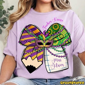 May include: A light purple t-shirt featuring a colorful Mardi Gras-themed design. The design includes a bow with purple and gold stripes, green and gold circles, a mask, and a pencil. The text "Teacher Krewe" and "Miss Oliver" are also included.