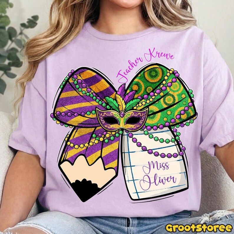 May include: A light purple t-shirt featuring a colorful Mardi Gras-themed design. The design includes a bow with purple and gold stripes, green and gold circles, a mask, and a pencil. The text "Teacher Krewe" and "Miss Oliver" are also included.