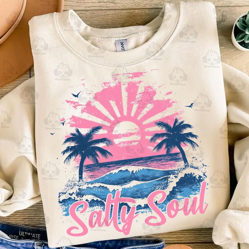 May include: A cream colored sweatshirt with a pink and blue graphic design of a sunset over the ocean with palm trees and the text "Salty Soul".