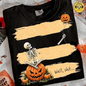 Könnte beinhalten: Ein schwarzes T-Shirt mit drei horizontalen, orangefarbenen Pinselstrich-Streifen. Ein Skelett sitzt auf einem großen Kürbis, während ein Skelettarm eine Schnur mit einem schwebenden Kürbis hält. Der Text "Well, shit..." ist in Schwarz aufgedruckt.
