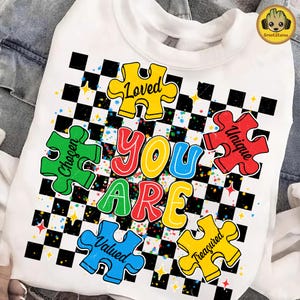 Puede incluir: Sudadera blanca con un diseño gráfico colorido. El diseño presenta las palabras "YOU ARE" en letras grandes y multicolores, rodeadas de piezas de rompecabezas con palabras como "Loved" y "Unique". Un patrón de cuadros blancos y negros llena el fondo.