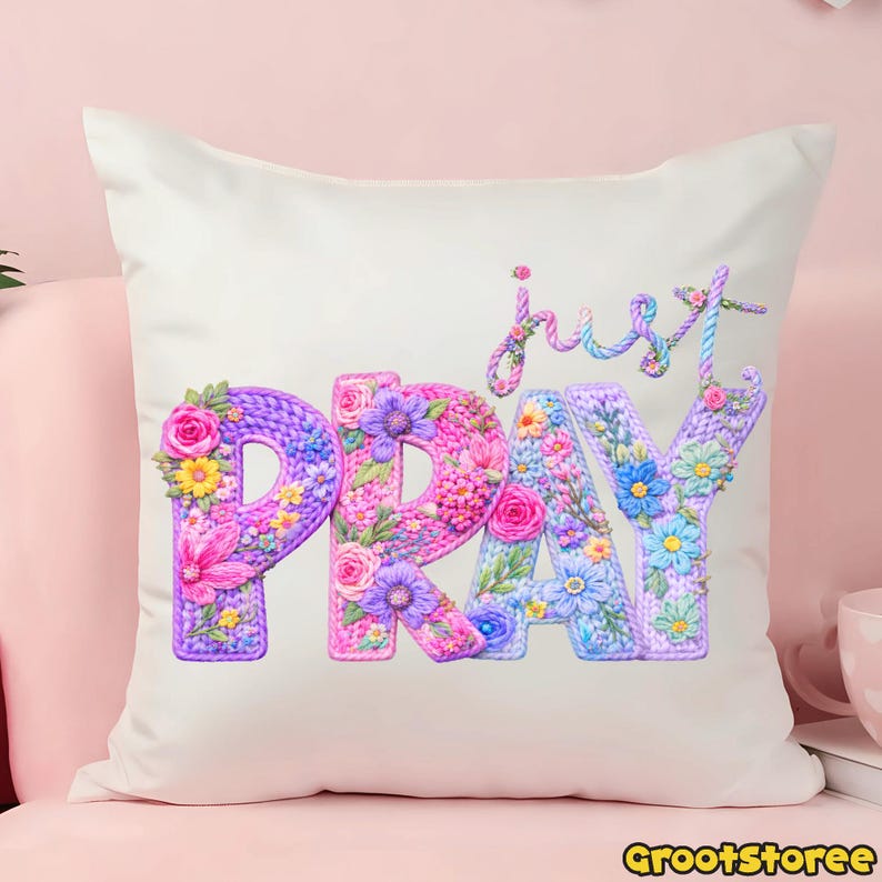 May include: A white square pillow with the words "just PRAY" embroidered in a floral design. The letters are in shades of purple, pink, and blue, with flower accents. The pillow is on a pink surface.
