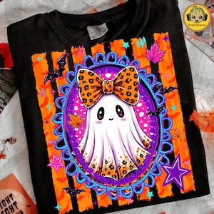 May include: A black t-shirt featuring a central graphic of a white ghost with a leopard print bow, framed by a blue ornate oval border. The background includes orange vertical stripes, black bats, pink stars, and colorful autumn leaves.