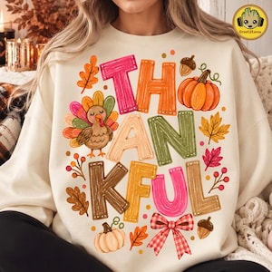May include: A cream-colored sweatshirt featuring a colorful graphic print of the word THANKFUL in block letters. The design includes a cartoon turkey, orange pumpkins, autumn leaves in red, yellow, and brown, acorns, and a red plaid bow.