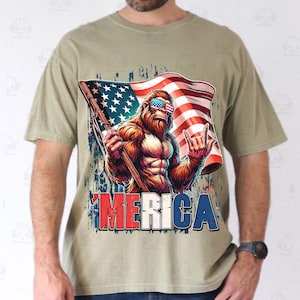 May include: A tan t-shirt featuring a graphic of a Bigfoot character wearing sunglasses with an American flag design. The Bigfoot is holding a flag and the word 'MERICA is printed below in red, white, and blue.
