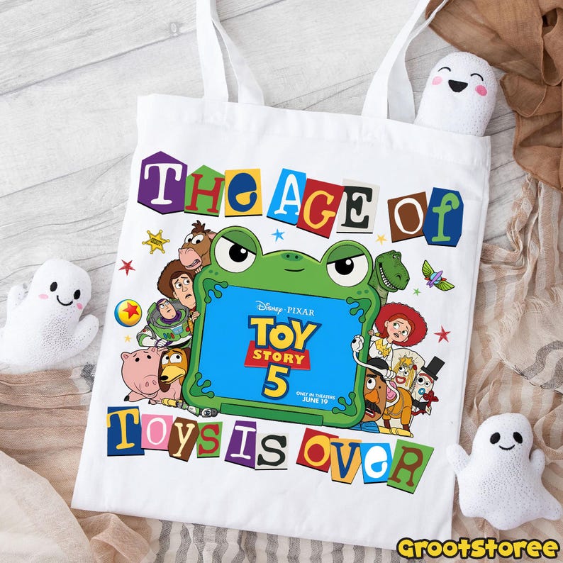 May include: A white tote bag featuring a colorful graphic of Disney Pixar Toy Story characters surrounding a green frog-shaped frame. The text reads The Age of Toy Story 5 Toys is Over, with Only in Theaters June 19 printed in the center.
