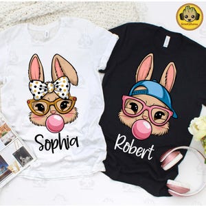 May include: Two white and black t-shirts with a cartoon bunny design. The white shirt has a pink bow and says "Sophia" and the black shirt has a blue baseball cap and says "Robert".