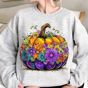 May include: A light gray sweatshirt featuring a vibrant, artistic pumpkin design. The pumpkin is a mix of orange, yellow, and purple, adorned with colorful flowers in shades of blue, purple, orange, and green. The design has a whimsical, hand-drawn style.