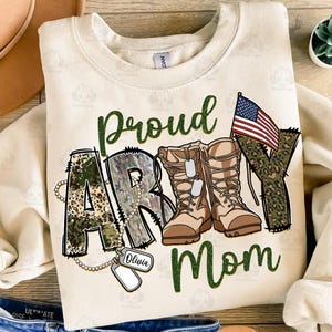 May include: A beige sweatshirt with the words "Proud Army Mom" in green and camouflage print. The word "Army" is made up of camouflage boots and a small American flag. There is a dog tag with the name "Olivia" on it.