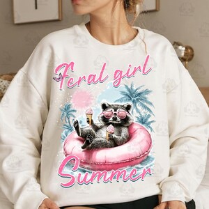 May include: White sweatshirt with a pink raccoon wearing sunglasses and holding an ice cream cone. The text "Feral girl summer" is printed on the front of the sweatshirt.