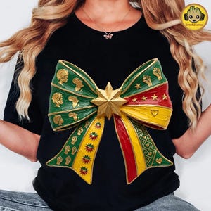 May include: Black t-shirt featuring a large, colorful bow design. The bow is green, yellow, and red with gold accents, including stars, fists, and African continent silhouettes. A gold star is at the center.