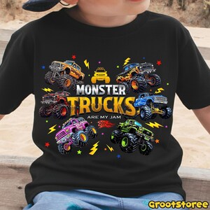 May include: Black t-shirt featuring a colorful graphic of monster trucks. The design includes various monster trucks in orange, blue, green, and pink, with the text "MONSTER TRUCKS ARE MY JAM". The shirt has a crew neck and short sleeves.