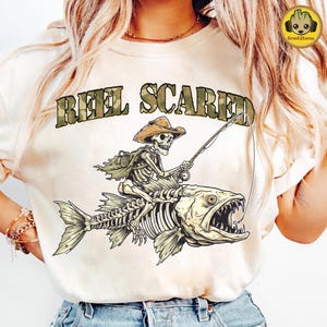 May include: Cream-colored t-shirt featuring a skeletal cowboy riding a skeletal fish, holding a fishing rod. The text "REEL SCARED" is printed above the image in camouflage-style lettering. The design is a humorous take on fishing.