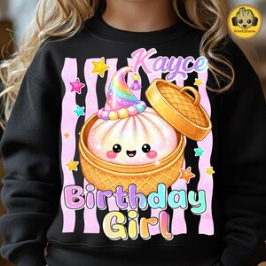 May include: A black sweatshirt featuring a graphic of a smiling steamed bun in a bamboo steamer basket, wearing a rainbow party hat. The design includes the text "Kayce" and "Birthday Girl" surrounded by pink vertical stripes and colourful stars.