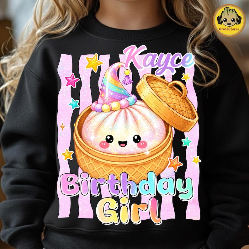 May include: A black sweatshirt featuring a graphic of a smiling steamed bun in a bamboo steamer basket, wearing a rainbow party hat. The design includes the text "Kayce" and "Birthday Girl" surrounded by pink vertical stripes and colourful stars.