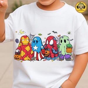 May include: A white t-shirt featuring a graphic print of four characters dressed as ghosts for Halloween. The characters resemble Iron Man, Captain America, Spider-Man, and Hulk, each holding a jack-o'-lantern candy bucket.