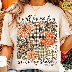 Puede incluir: Una camiseta color crema con un estampado gráfico de una cruz a cuadros blancos y negros, calabazas con patrones de leopardo y lunares, y flores de cuadros naranjas. El texto dice I will praise him in every season PSALM 146:2.