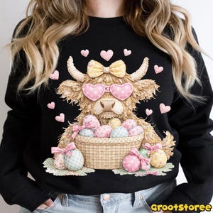 Puede incluir: Sudadera negra con un dise&ntilde;o de vaca de las Tierras Altas con gafas de sol en forma de coraz&oacute;n y un lazo. La vaca sostiene una cesta de huevos de Pascua. Corazones y lazos rosas decoran el dise&ntilde;o. El texto "GrootStoree" est&aacute; en la parte inferior derecha.