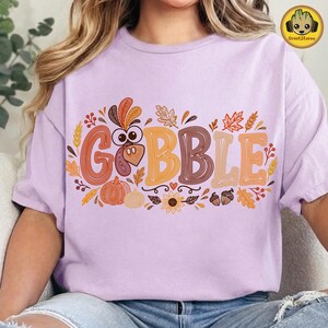 May include: A light purple t-shirt featuring the word GOBBLE in large, multi-colored, line-art style letters. The design includes a turkey head, pumpkins, sunflowers, acorns, and autumn leaves in shades of orange, brown, and yellow.