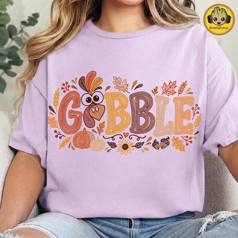 May include: A light purple t-shirt featuring the word GOBBLE in large, multi-colored, line-art style letters. The design includes a turkey head, pumpkins, sunflowers, acorns, and autumn leaves in shades of orange, brown, and yellow.