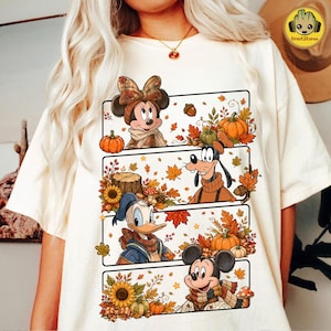 May include: A cream-colored t-shirt featuring a graphic print of four horizontal panels. Each panel displays a Disney character, including Minnie Mouse, Goofy, Donald Duck, and Mickey Mouse, surrounded by autumn leaves, pumpkins, and sunflowers.