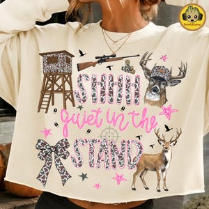 May include: A cream-coloured sweatshirt featuring a graphic print of a hunting stand, a rifle, two deer, a bow, and the text "SHHH quiet in the STAND". The design includes cow print patterns, pink stars, and silhouettes of birds against a light background.