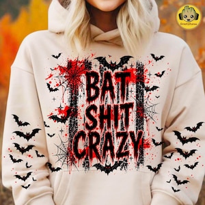 May include: A beige hooded sweatshirt featuring a graphic print with the text BAT SHIT CRAZY in black distressed lettering. The design includes black bat silhouettes, spider webs, and red paint splatter accents across the front and sleeves.