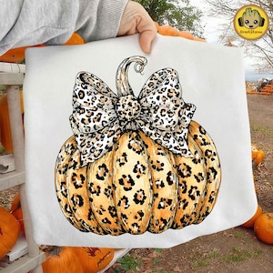 May include: A decorative pumpkin with a leopard print design and a matching bow. The pumpkin is orange with black spots, and the bow features a similar pattern. The image is set against a background of pumpkins.