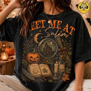 May include: A black acid-wash t-shirt featuring a graphic print with the text "MEET ME AT Salem". The design includes a jack-o'-lantern, a crystal ball with a house inside, an open book titled "WITCHES COMPENDIUM", a broom, and a candle.