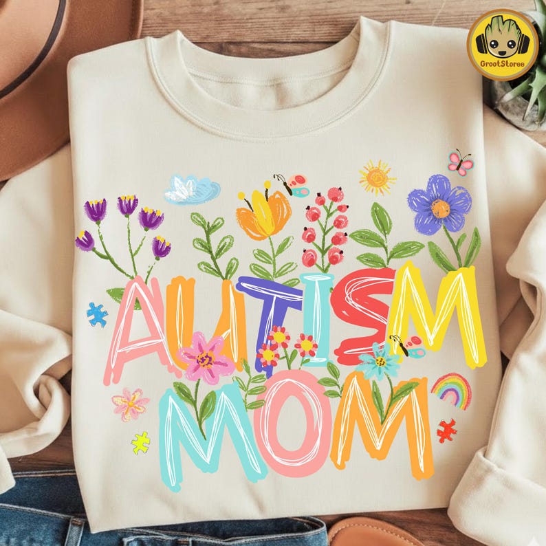 Puede incluir: Sudadera color crema con las palabras "AUTISM MOM" en letras coloridas y divertidas. El dise&ntilde;o incluye ilustraciones florales y de mariposas, con un arco&iacute;ris y un sol. La sudadera es de estilo casual y c&oacute;modo.