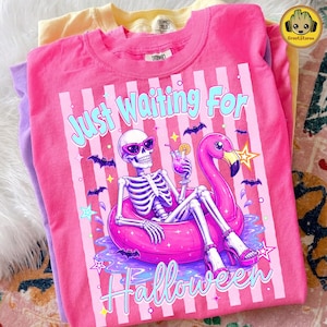 Könnte beinhalten: Ein leuchtend pinkes T-Shirt mit einer Grafik eines Skeletts, das eine pinke Sonnenbrille und High Heels trägt, in einem pinken Flamingo-Schwimmring entspannt und einen Drink hält. Der Text lautet Just Waiting For Halloween mit Fledermäusen.