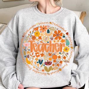 May include: A light gray sweatshirt with a circular floral design. The design features the word "Teacher" in orange, surrounded by flowers, pumpkins, and the words "Motivator, Warm, Dedicated, Loving, Caring, Fun, Kind, Reliable, Fair, Compassionate."