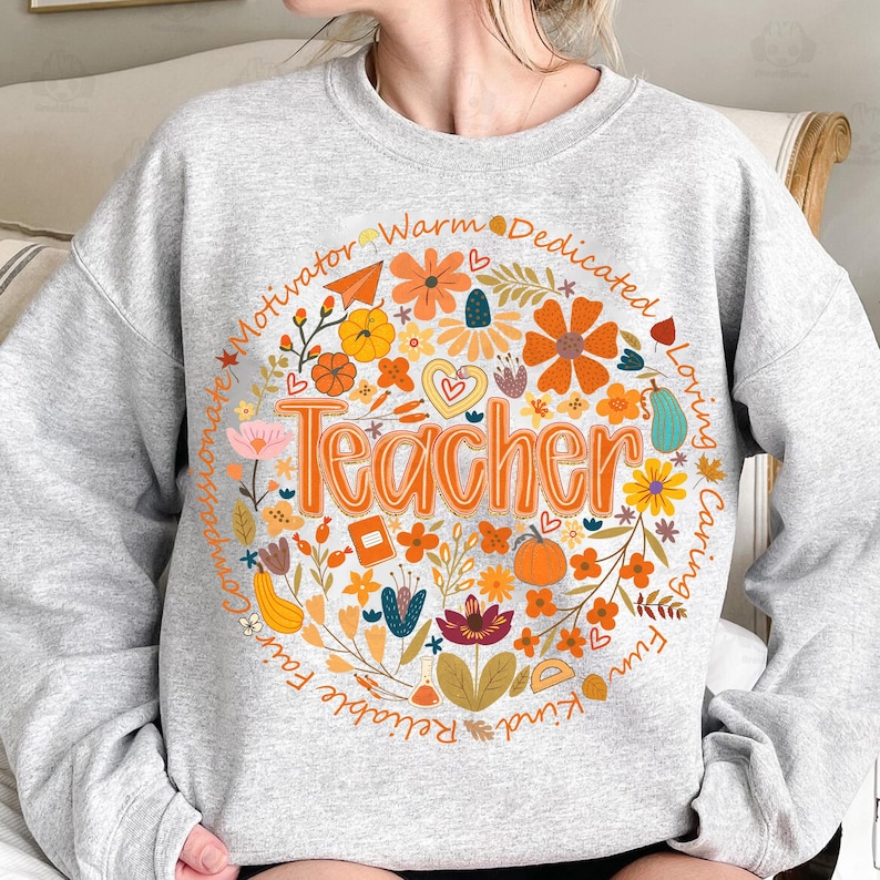 May include: A light gray sweatshirt with a circular floral design. The design features the word "Teacher" in orange, surrounded by flowers, pumpkins, and the words "Motivator, Warm, Dedicated, Loving, Caring, Fun, Kind, Reliable, Fair, Compassionate."