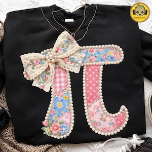 Puede incluir: Sudadera negra con un gran símbolo Pi decorativo. El símbolo está adornado con patchwork floral, lunares rosas y un lazo de encaje. Los bordes están delineados con perlas de imitación. Un collar dorado está drapeado sobre la sudadera.