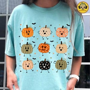 May include: A light blue t-shirt featuring a graphic print of nine cartoon pumpkins with various facial expressions and patterns, including polka dots, hearts, and stars, interspersed with small black bats and orange stars across the front.