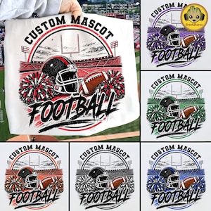 Puede incluir: Una camiseta blanca con un gráfico de un estadio de fútbol americano que incluye un casco, un balón y pompones. El texto CUSTOM MASCOT FOOTBALL está impreso en negro. El diseño se muestra en varias variantes de color como rojo, morado, verde y azul.