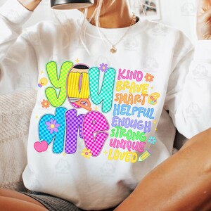 Op de afbeelding: Witte sweatshirt met de woorden "You are" in grote, kleurrijke, bubbelletters. De woorden "Kind, Brave, Smart, Helpful, Enough, Strong, Unique, Loved" staan ook op de trui. Het ontwerp bevat bloemen, een potlood en een gloeilamp.
