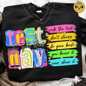 May include: Black sweatshirt with colorful text that reads "Test, you got this, day." Additional text includes "rock the test, don't stress, do your best, you know it, now show it." The design features various school-themed graphics.