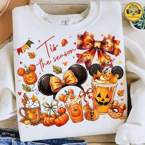 May include: A white sweatshirt featuring a colorful graphic with the text 'Tis the season'. The design includes orange pumpkin-shaped Mickey Mouse ears, a large patterned bow, a jack-o'-lantern drink, a mug with marshmallows, and autumn leaves.